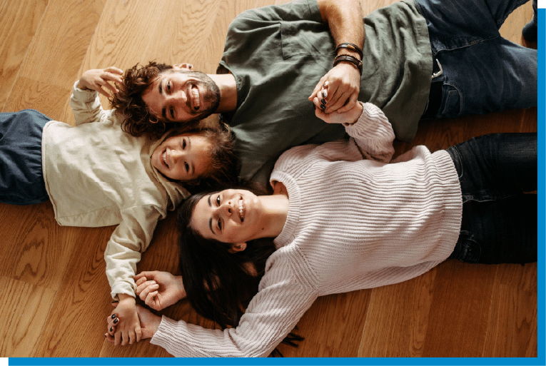 Family lying on the living room floor smiling and holding hands during a playful moment