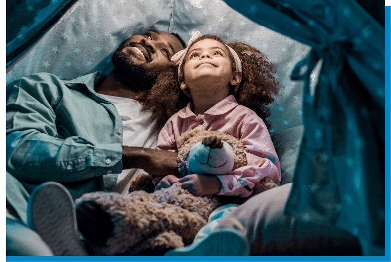 Father and daughter smiling together in a cozy nighttime fort, holding a stuffed bear